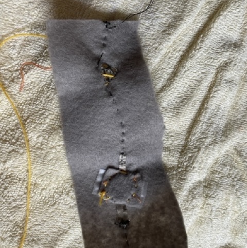 finishing up the conductivity tester by just sowing down on top of the tape to keep the battery in place and not messing with it any further. The light was finicky because the needle was too thick to actually go through the holes in the light so i had to sow around it to pin it in place. I was going to use yellow thread to hold it down more securely but that ended up just interfering with the connection so I removed it.